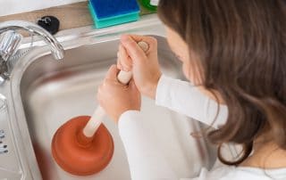Woman Using Plunger In Kitchen Sink At Home
