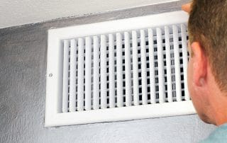 Mature male peering inside an upper wall white grid air duct on a silver wall near a white ceiling